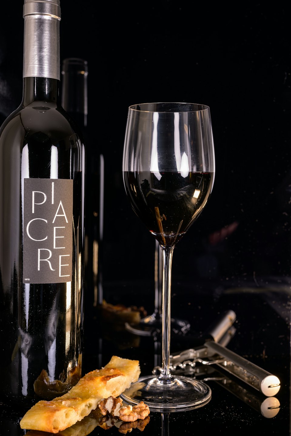 Bottiglia di vino PIACERE con bicchiere e noci su una superficie riflettente.